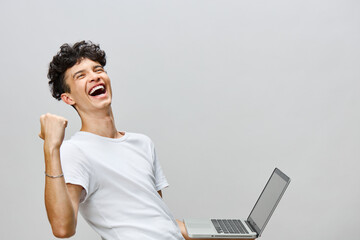 joyful young man celebrating success while holding a laptop, light gray background, casual attire,...