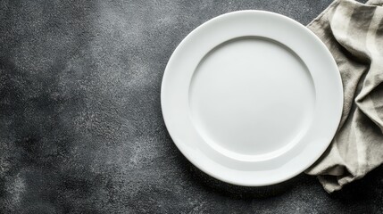 A simple white plate placed on a dark textured surface with a striped cloth nearby, ideal for culinary presentations.