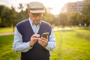 Fototapeta premium Portrait of happy senior man in park on sunny day. He is messaging on smartphone.
