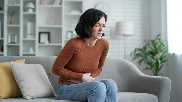 Young adult female suffering from abdominal pain sitting on sofa in living room at home. Sad woman keeps her hands on her stomach. She has poisoning, constipation, bloating, spasms or menstruation
