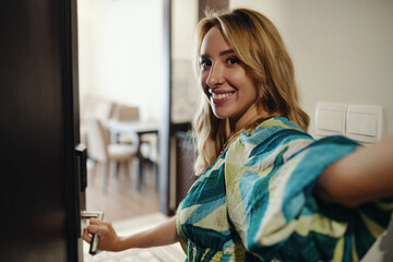 Woman with blonde hair smiling and holding door handle while looking back. Indoor setting with dining area in background showing an inviting atmosphere