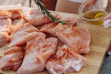 Close-up of a woman's hands adding spice marinade, paprika and olive oil with fresh rosemary to meat steaks for cooking on a home patio grill