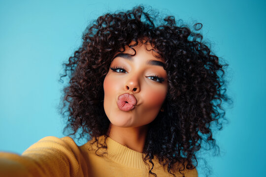 A playful portrait of a woman with curly hair making a kissy face while taking a selfie against a bright blue background