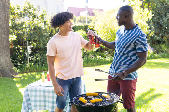 Grilling food and toasting with drinks, two diverse male friends enjoying outdoor barbecue