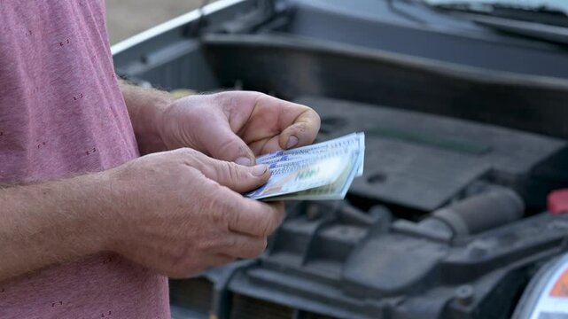 Payment to mechanic for repairing of car. Caucasian man counting dollar bills and giving them to car service worker in front of open hood of a car