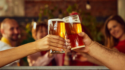 Close-up of two raised glasses filled with amber beer, with blurry background action suggesting lively sports crowd enjoying game together. Concept of Oktoberfest, brewery, vacation, Friday mood.