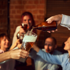 As bottle pours foamy beer into glass, group of people in background cheer, raising their drinks. Setting feels like pub or bar filled with happy energy. Concept of Oktoberfest, brewery, Friday mood.