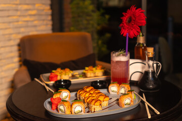 Set of various delicious mouth-watering sushi rolls on the table with a cocktail, soy sauce, chopsticks and a vase with a flower in the restaurant in the evening, a selective focus.