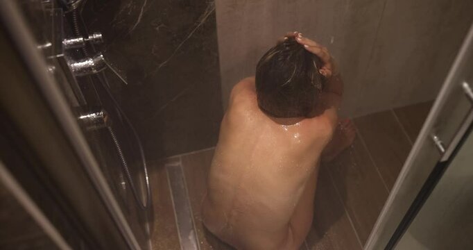 Female person sits in shower stall under running water in despair in modern bathroom of accommodation. Sadness and disappointment backside view