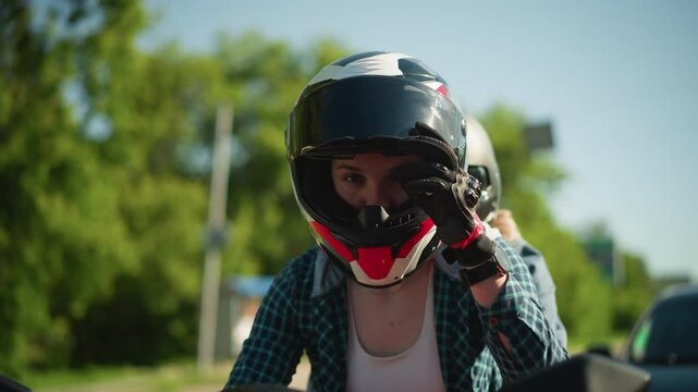 Two friends ride a red power bike, both wearing helmets, with the rider closing her visor, the rider wears black gloves and a wristwatch, a car is visible behind them