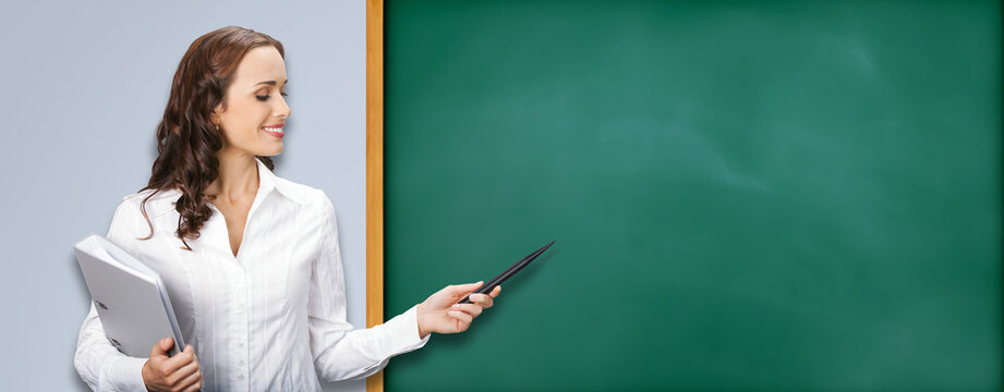 Happy smiling young beautiful business woman or teacher showing pointing with pointer at green chalkboard with sign text space, isolated on grey gray wall background. Mockup image.