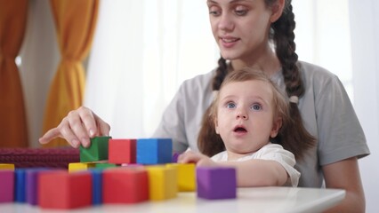 Obraz premium mother plays with her child. happy childhood dream concept for a little child. a small child sits at the white table on his mother lifestyle lap and plays with beautiful colorful cubes