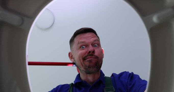 Funny plumber opens toilet bowl and scratches nape with plunger in restroom. Emotional man cleans clog in pipe with traditional tool view from bottom