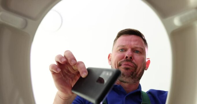 Plumber in uniform opens toilet bowl and takes out dropped mobile phone in restroom. Disgusted man finds smartphone in john in public lavatory
