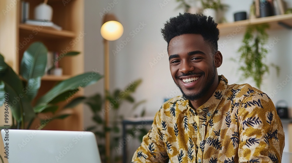 Wall mural A smiling man in a patterned shirt sits at a desk with a laptop, surrounded by plants and a cozy interior.