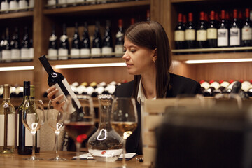 Experienced caucasian female sommelier in classic clothes sitting at table during preparing to wine tasting on various bottles background shelf. Sommelier exam to study different wine. 