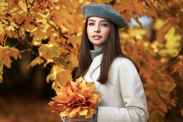 Autumn portrait of a beautiful happy smiling woman with yellow maple leaves in a gray beret in the park