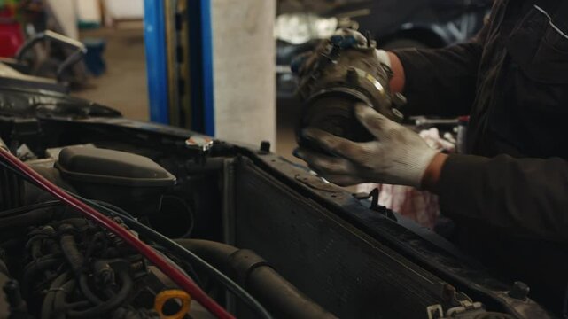 A car mechanic in uniform takes out a car part AC air conditioning compressor from under the hood