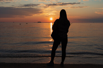 Silhouette of a young girl with a backpack on the beach during sunset in summer, people swimming in the sea and a ferry sailing, photo view from the back.