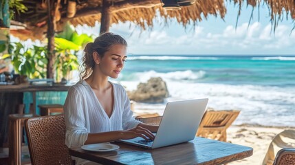 A woman works on her laptop at a beachside table, surrounded by tropical scenery and ocean waves, enjoying a serene remote working experience.
