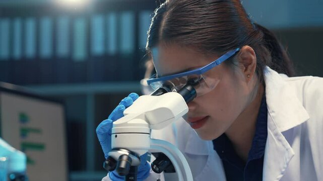 Scientist wearing a lab coat and safety glasses is carefully examining a sample through a microscope in a modern laboratory setting