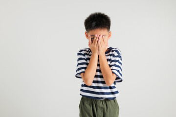 Asian portrait young kid boy in depressed bad mood covering face with hands and peering out with one eye between fingers studio shot isolated on white background, primary child surprised and shocked © sorapop