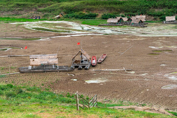 A dried-up lakebed with several wooden platforms and small huts built on it. Some boats are grounded on the cracked mud. In the background, there are hills covered in greenery.