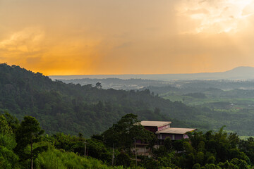 A panoramic view of a lush green valley bathed in the golden light of a setting sun. A small, two-story house nestled among the trees stands out against the dramatic sky.