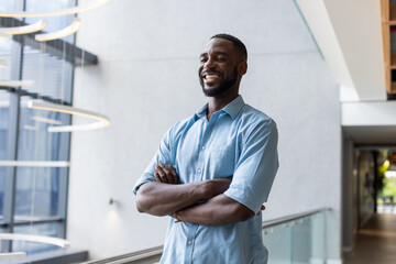 Smiling african american man in blue shirt standing with arms crossed in modern office