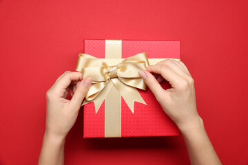 Woman decorating gift box with bow on red background, top view