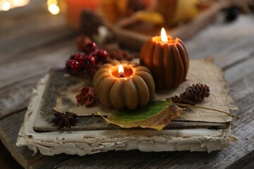 Burning candles in shape of pumpkins, old book and autumn decor on wooden table, closeup