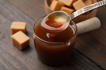 Taking tasty caramel sauce with spoon from glass measuring cup at wooden table, closeup
