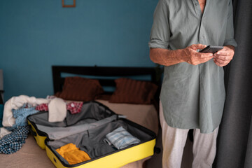 Senior man packing his luggage for upcoming trip, using the checklist on smartphone. Retired...