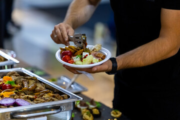 A person serves themselves a variety of grilled dishes from a buffet at a casual outdoor event during a sunny afternoon