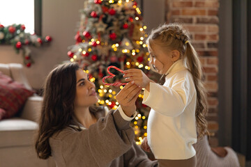 Little girl and her mom sitting near Christmas tree and holding heart-shaped candy on festive Christmas tree background. New year holidays, copy space, selective focus