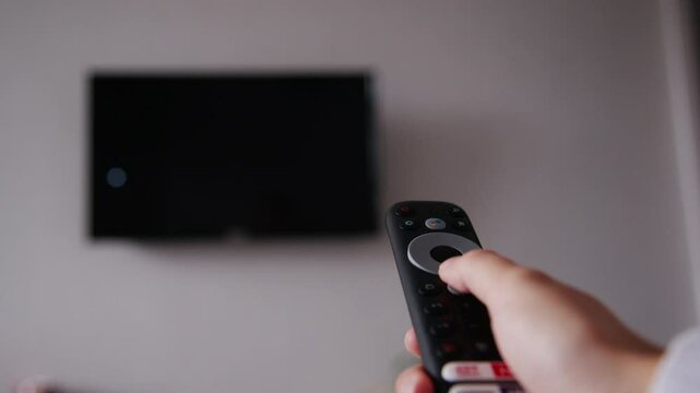 Close-up of a woman's hand holding a TV remote control and changing TV channels. In the background of the hand is a wall with a TV in out of focus. Blurred TV