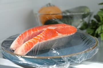 Plate of salmon with plastic food wrap on shelf in refrigerator, closeup