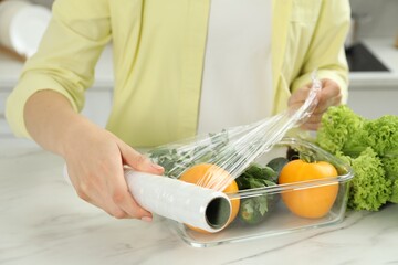 Woman putting plastic food wrap over glass container with fresh vegetables at white marble table,...