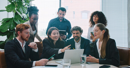 Group of people gathering around one laptop and listening to confident Asian woman. Female describing something with hand gestures while pointing at screen. Showing growth possibilities.