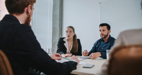 Beautiful Caucasian female sitting among colleagues during team building meeting. Woman with glasses and long hair wearing white shirt and black suit jacket. Hand gestures. Sales team discussing.