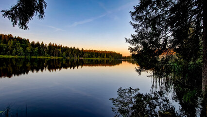 Nature reserve Pláničský pond in Černá v Pošumaví in the evening. The calm water mirrors the surrounding trees and sky. A bench and a small boat are visible by the shore. 