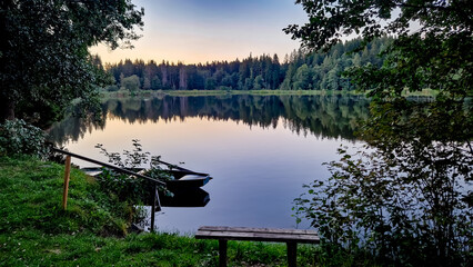 Nature reserve Pl&aacute;ničsk&yacute; pond in Čern&aacute; v Po&scaron;umav&iacute; in the evening. The calm water mirrors the surrounding trees and sky. A bench and a small boat are visible by the shore. 