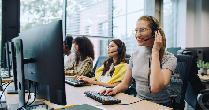 Pretty worker wearing headset during online consultation with her client. Enjoying providing quality services to customers. Beside woman sitting coworkers at their computers.