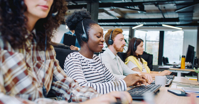 Professional multi-ethnic managers working in call center office. People sitting in row at desk and using computers. Discussing something with customers on headset. Team of workers answering calls. - Powered by Adobe