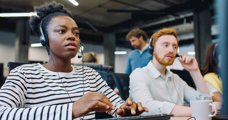 Multi-ethnic people sitting in row at their workplaces in office center. Hardworking managers or operators calling to customers while wearing headsets. Entering data on computers of conversation.