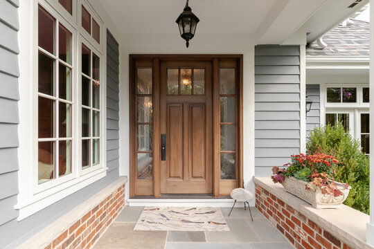 A front door and covered porch detail on a home with grey siding, white trim, red brick, a wooden front door.