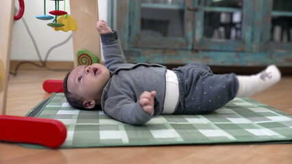 Baby lying on a checkered activity mat, gazing up at colorful toys with a curious expression....