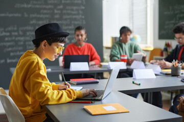 Side view portrait of teenage girl sitting at table and using computer during lesson in school classroom, copy space