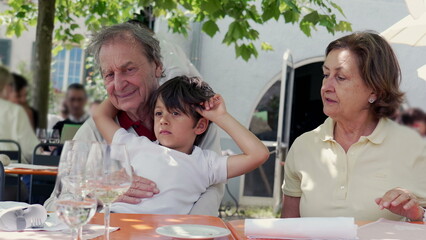 Grandfather lovingly embraces his young grandson at an outdoor cafe, with the grandmother watching...