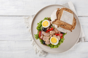 French Pan Bagnat sandwich with salad nicoise consisting of hard-boiled eggs, tuna, vegetables and seasoned with olive oil close-up on a plate on a wooden table. Horizontal top view from above
